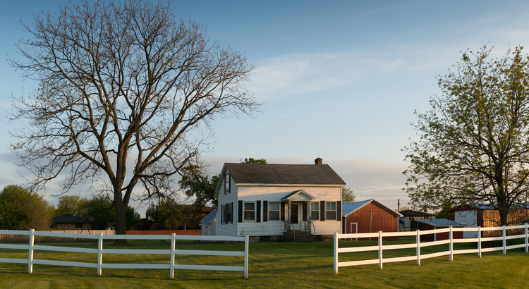 farm house with white fence and mature trees against a blue sky