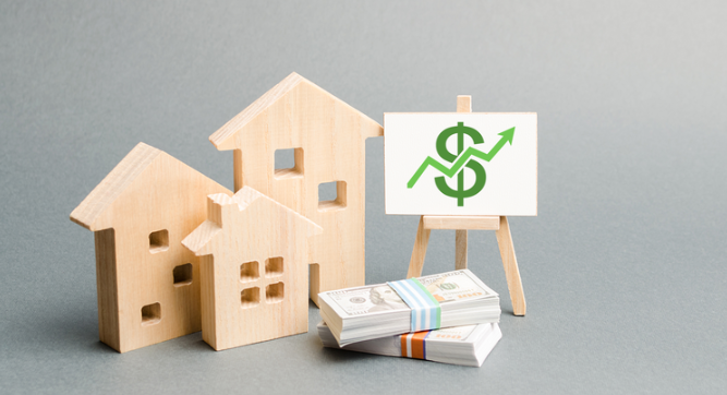 3 wooden toy houses, 2 stacks of hundred dollar bills and a toy easel displaying a green dollar sign with graph arrow trending up set against a grey background
